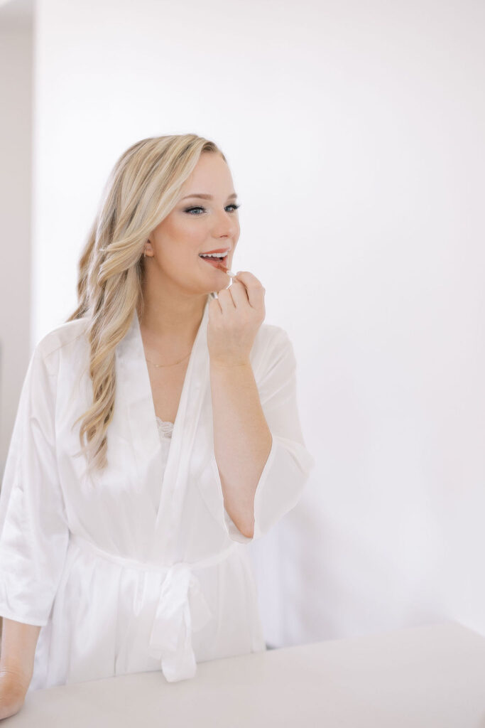 A beautiful bride wearing a satin robe and putting on lipstick on her wedding day.  Need help with stress-free wedding planning? Talk to Moments by Maddie!