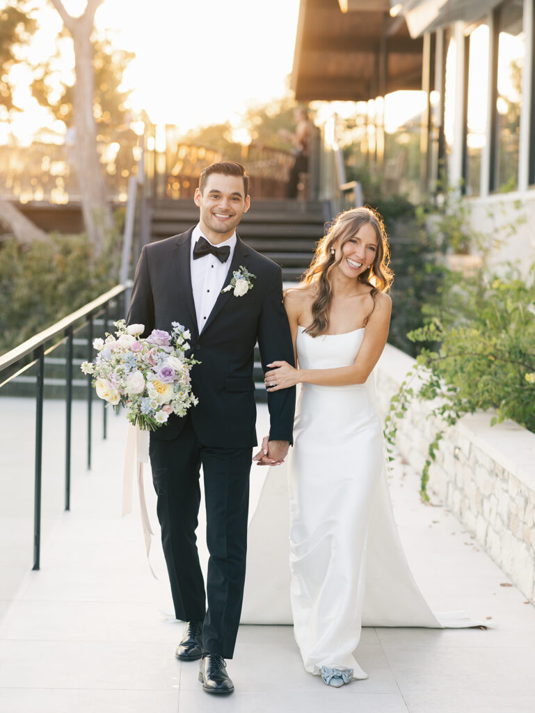 Bride and groom walking hand in hand with joyful smiles and a pastel bridal bouquet, capturing the calm, coordinated energy of a well-planned wedding day.