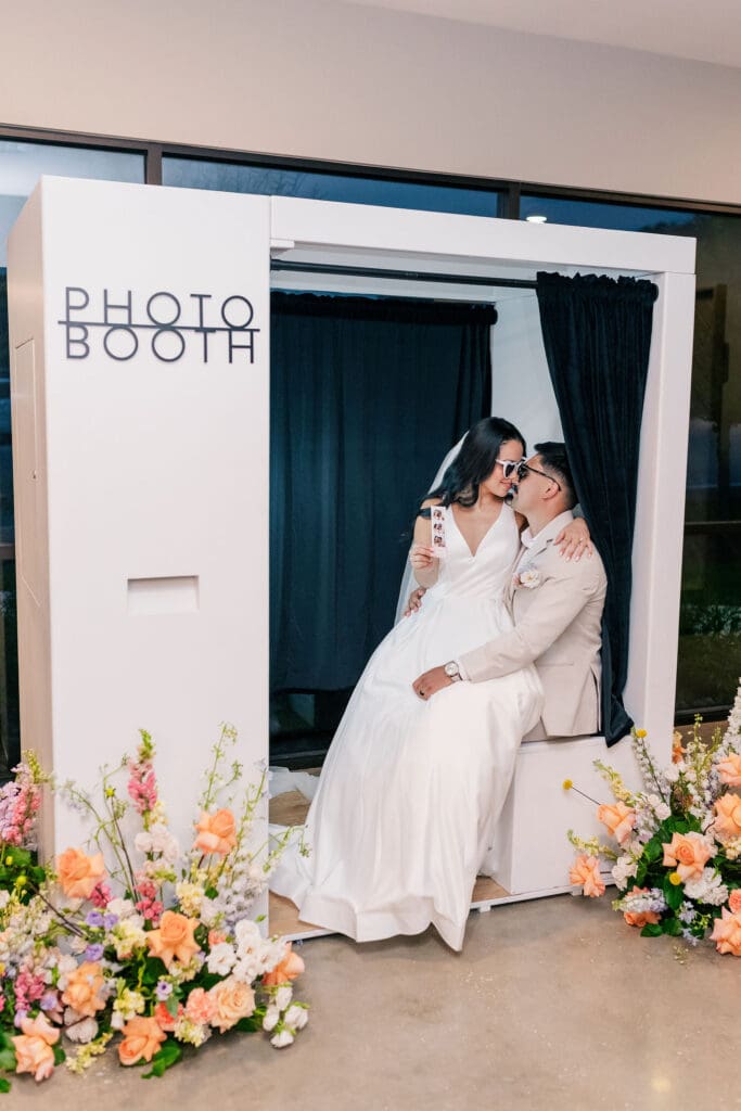 Modern white photo booth with black curtain and floral styling, featuring a newlywed couple posing—an interactive wedding design idea on a budget from Austin wedding planner Moments by Maddie.