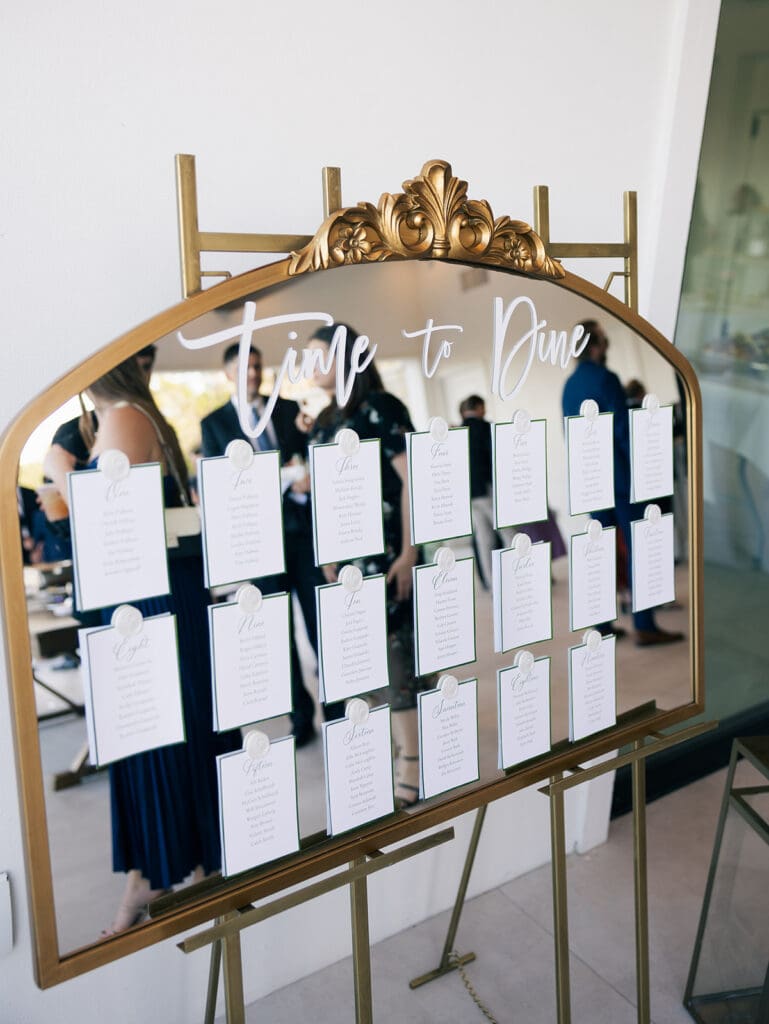 Mirror escort card display with gold frame and acrylic “Time to Dine” sign, a stylish wedding design idea on a budget that doubles as decor.