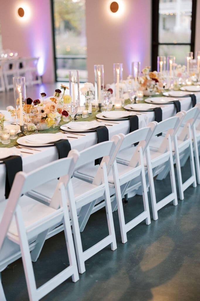 Long reception table with black napkins, neutral florals, and glowing taper candles—an elegant wedding design idea on a budget with major visual impact.