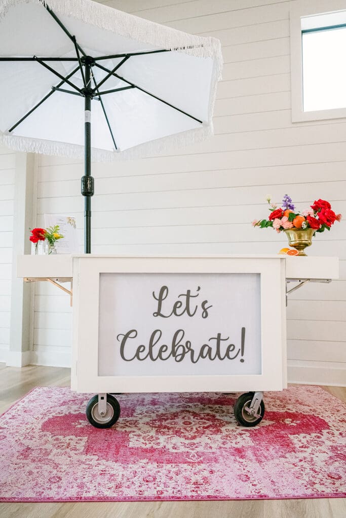 A white bar cart with a “Let’s Celebrate!” sign and colorful flowers, styled with a fringe umbrella and pink rug for a playful cocktail hour setup.