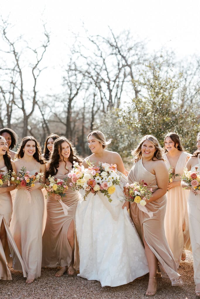 Bride surrounded by bridesmaids in champagne gowns holding vibrant bouquets
