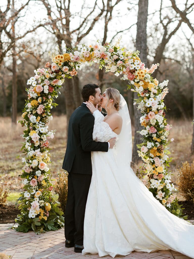 Bride and groom kissing under a vibrant floral arch at a fall wedding in Texas