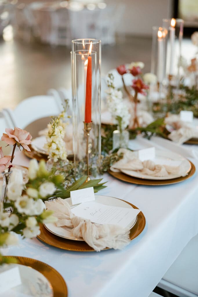 Reception table with orange taper candles, gold chargers, and pastel floral arrangements